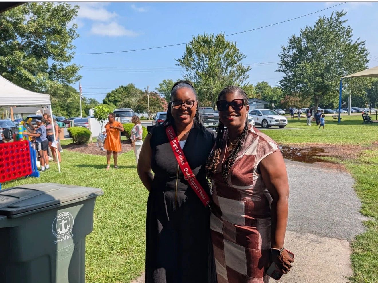 Two women at an outdoor community event, one wearing a red sash
